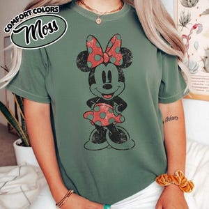 May include: A moss green t-shirt with a distressed Minnie Mouse graphic. The Minnie Mouse design is black with a red polka dot bow and dress. The shirt features the words "COMFORT COLORS" and "Moss" in a circular design.