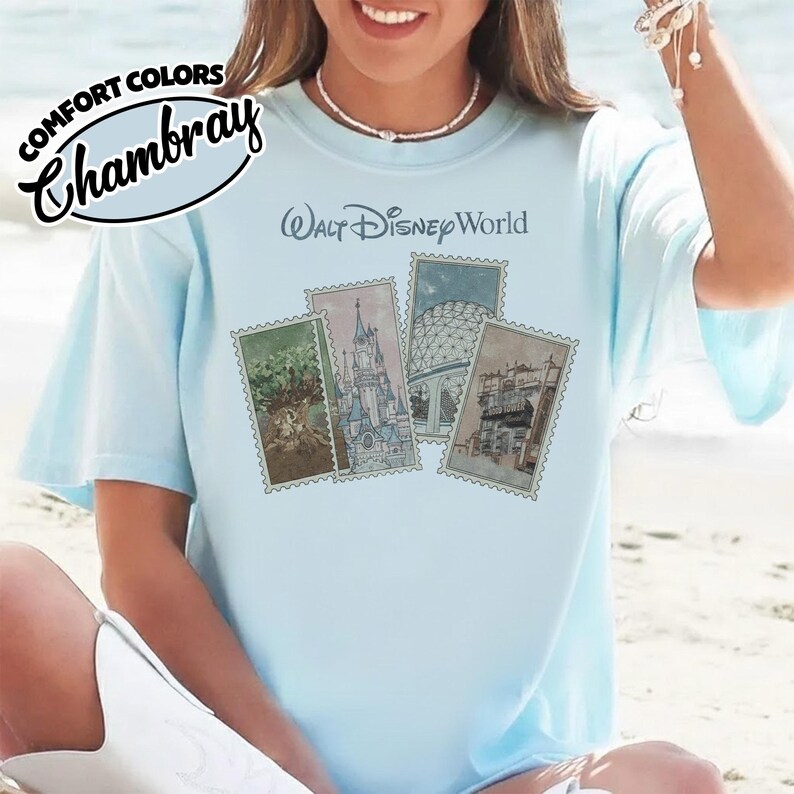 Op de afbeelding: Lichtblauw T-shirt met "Walt Disney World" gedrukt boven vier postzegelontwerpen. De postzegels tonen illustraties van Disney park bezienswaardigheden. Het shirt heeft ook de woorden "Comfort Colors Chambray" linksboven gedrukt.