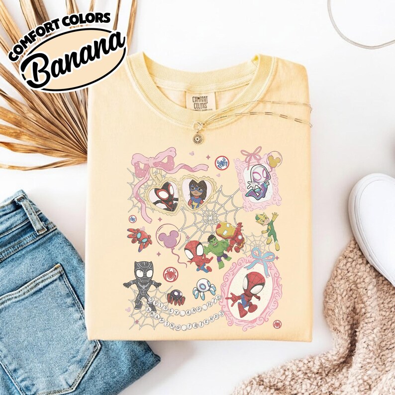 K&ouml;nnte beinhalten: Hellgelbes Comfort Colors T-Shirt mit einer Cartoon-Grafik mit verschiedenen Spider-Man-Charakteren. Das Shirt hat den Text "Comfort Colors Banana" in einem schwarz-wei&szlig;en Oval. Die Grafik enth&auml;lt rosa Rahmen und Spinnweben.