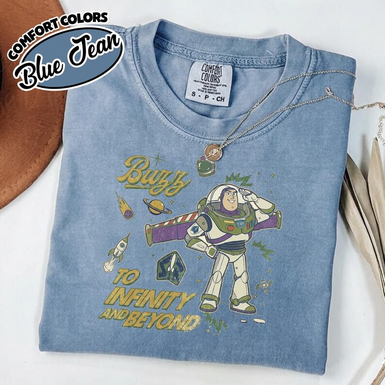 May include: A blue jean colored t-shirt featuring a graphic of Buzz Lightyear saluting, surrounded by space-themed icons including a rocket, planet, and comet. The text reads Buzz TO INFINITY AND BEYOND. The shirt has a Comfort Colors brand label.