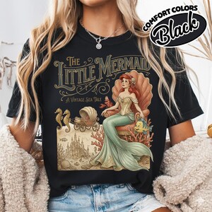 以下が含まれることがあります： 黒いTシャツ。ヴィンテージスタイルのリトルマーメイドのイラストが特徴です。デザインには、貝殻に座るアリエル、海の馬車、そして「A Vintage Sea Tale」というテキストが含まれています。「Comfort Colors」のブランドです。