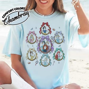 May include: Light blue t-shirt with a graphic featuring cartoon princesses in ornate frames. The shirt has the words "Comfort Colors Chambray" printed on the upper left side. The princesses are in oval frames with bows and crowns.
