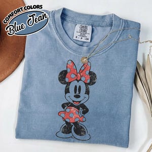 May include: Blue jean Comfort Colors t-shirt with a distressed Minnie Mouse graphic. The graphic shows Minnie Mouse wearing a red polka dot bow and dress. A gold necklace is draped across the shirt. The shirt has the Comfort Colors brand tag.