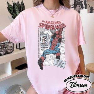 K&ouml;nnte beinhalten: Hellrosa T-Shirt mit Comic-Grafik von Spider-Man. Der Aufdruck enth&auml;lt den Text "the AMAZING SPIDER-MAN" und "COMFORT COLORS Blossom". Das T-Shirt hat einen Rundhalsausschnitt.