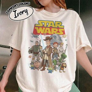 May include: Ivory-colored t-shirt featuring a graphic that combines Star Wars and Toy Story characters. The design includes Woody, Buzz Lightyear, and other characters, with the Star Wars logo at the top. The shirt has a Comfort Colors label.