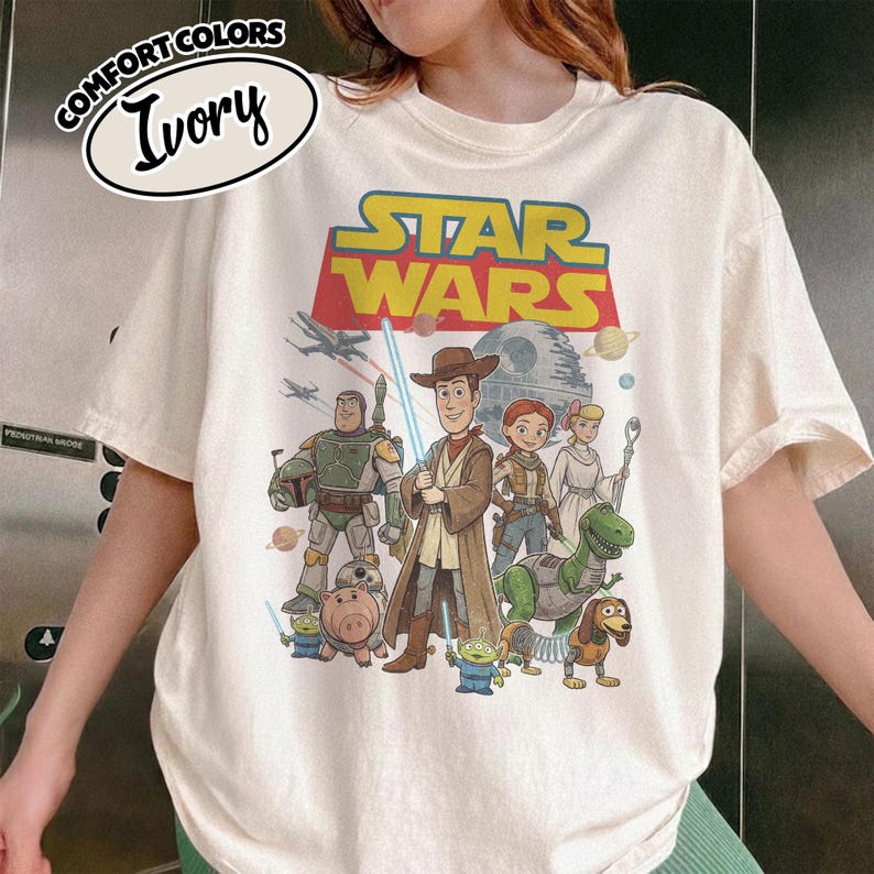 May include: Ivory-colored t-shirt featuring a graphic that combines Star Wars and Toy Story characters. The design includes Woody, Buzz Lightyear, and other characters, with the Star Wars logo at the top. The shirt has a Comfort Colors label.