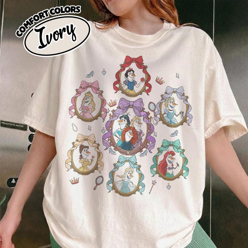 May include: Ivory-colored t-shirt featuring framed portraits of animated characters. The portraits are surrounded by decorative bows and crowns. The shirt has the words "COMFORT COLORS" and "Ivory" printed on the top left.