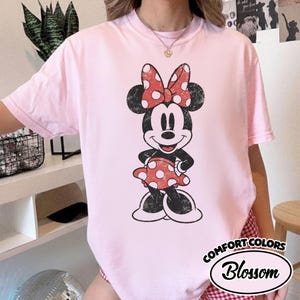 May include: A pale pink Comfort Colors t-shirt with a distressed Minnie Mouse graphic. Minnie wears a red and white polka dot bow and dress. The shirt has the text "Comfort Colors Blossom" in a white oval.