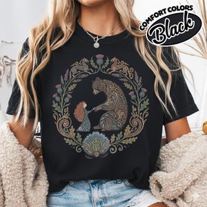 May include: Black t-shirt with a circular design featuring a bear and a girl holding hands, surrounded by floral and animal motifs. The text "COMFORT COLORS Black" is printed on the upper right. The design is in shades of brown, green, and blue.