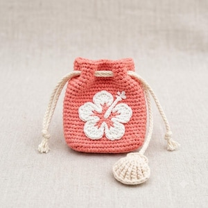 May include: Coral-colored crochet drawstring bag with a white hibiscus flower applique. The bag features a cream-colored drawstring and a small, cream-colored seashell charm. The bag is handmade and has a textured appearance.