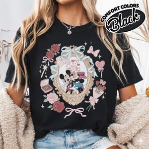 Puede incluir: Una camiseta negra con un estampado gr&aacute;fico de Mickey y Minnie Mouse bes&aacute;ndose dentro de un marco ovalado ornamentado. El dise&ntilde;o incluye rosas rosadas, un ramo, una caja de bombones con la etiqueta Be Mine, corazones y un lazo azul claro.