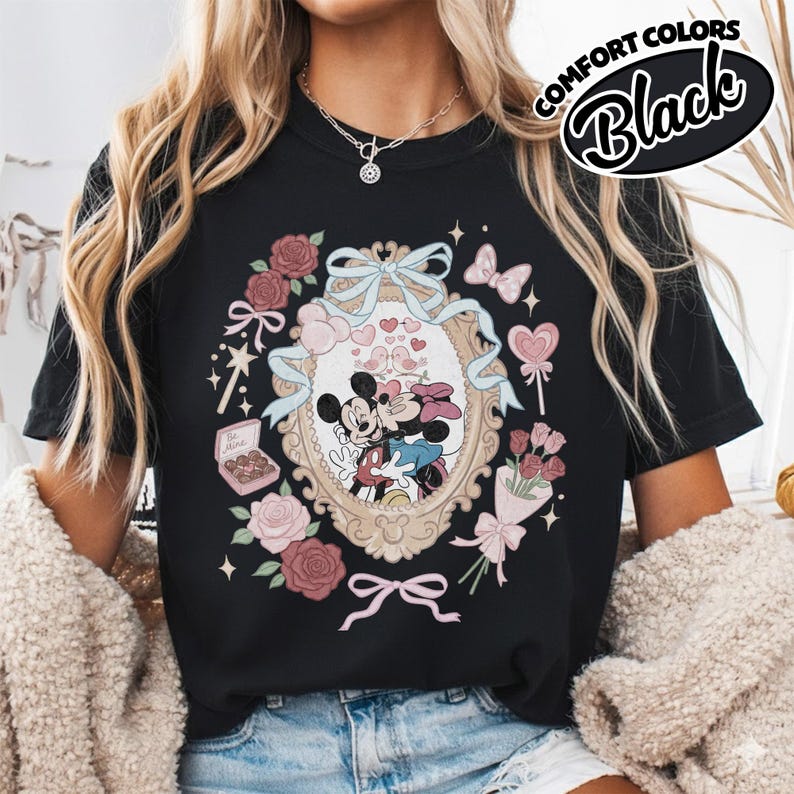 Puede incluir: Una camiseta negra con un estampado gr&aacute;fico de Mickey y Minnie Mouse bes&aacute;ndose dentro de un marco ovalado ornamentado. El dise&ntilde;o incluye rosas rosadas, un ramo, una caja de bombones con la etiqueta Be Mine, corazones y un lazo azul claro.