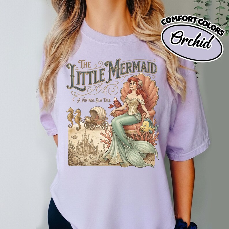 以下が含まれることがあります： オーキッドコンフォートカラーズのTシャツ。ヴィンテージイラストの「リトルマーメイド」が描かれています。イラストには、貝殻の玉座に座るアリエル、タツノオトシゴ、馬車、お城が描かれています。「A Vintage Sea Tale」の文字も含まれています。