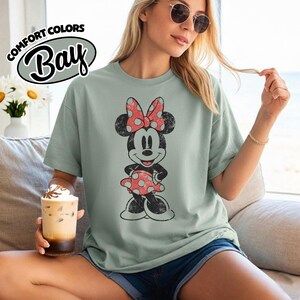 May include: A pale green t-shirt with a distressed Minnie Mouse graphic, sporting a red polka dot bow and dress. The shirt features the text "COMFORT COLORS Bay" in a circular design on the top left.