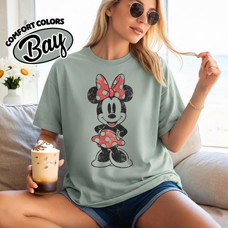May include: A pale green t-shirt with a distressed Minnie Mouse graphic, sporting a red polka dot bow and dress. The shirt features the text "COMFORT COLORS Bay" in a circular design on the top left.