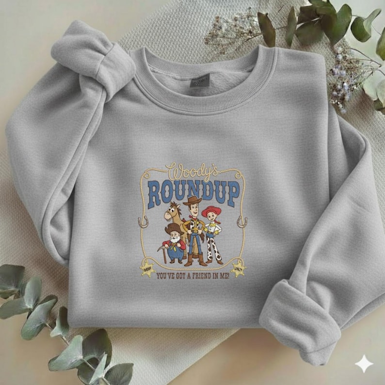 Puede incluir: Una sudadera gris claro de cuello redondo con un estampado gr&aacute;fico central titulado Woody's Roundup. El dise&ntilde;o incluye a los personajes Woody, Jessie, Bullseye y Stinky Pete enmarcados por un borde de cuerda con el texto You've Got A Friend In Me.