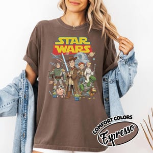 May include: Brown t-shirt featuring a Star Wars graphic with characters from the movie. The design includes the Star Wars logo in yellow and red, and a group of characters from the movie. The shirt also has the words "Comfort Colors Espresso" printed on it.