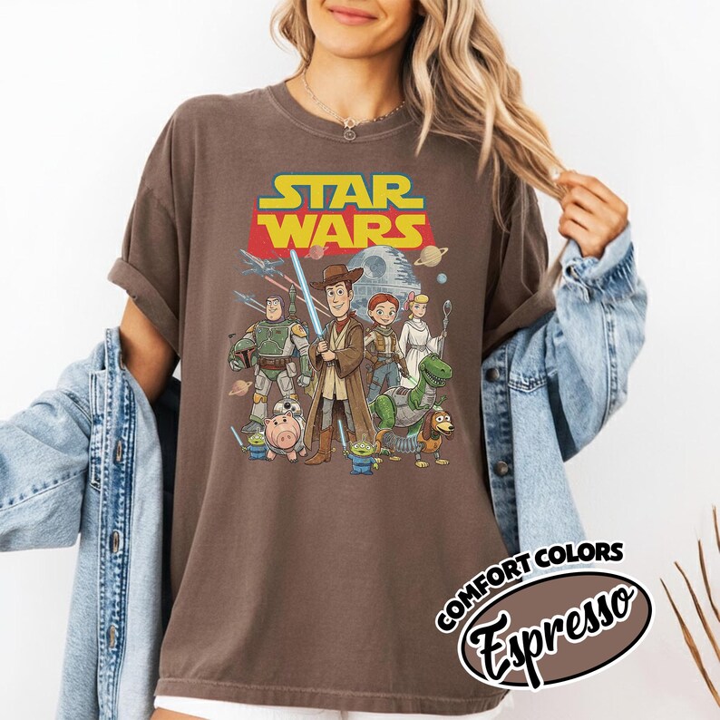 May include: Brown t-shirt featuring a Star Wars graphic with characters from the movie. The design includes the Star Wars logo in yellow and red, and a group of characters from the movie. The shirt also has the words "Comfort Colors Espresso" printed on it.