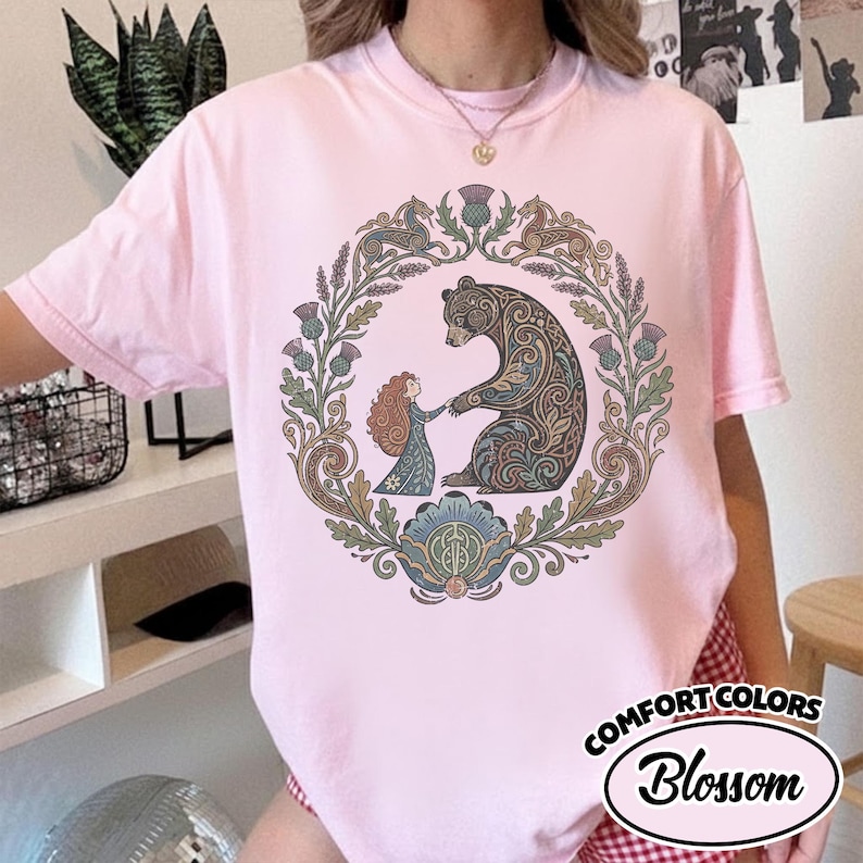 May include: Pink Comfort Colors t-shirt with a circular design featuring a bear and a person holding hands. The design is surrounded by a floral wreath. The text "Blossom" is in a white oval.
