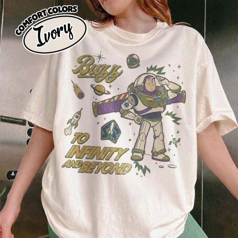 May include: An ivory short-sleeve t-shirt featuring a graphic of Buzz Lightyear saluting. The design includes space-themed elements like a rocket, planet, and stars, with the text "Buzz" and "To Infinity and Beyond" in a vintage-style font.