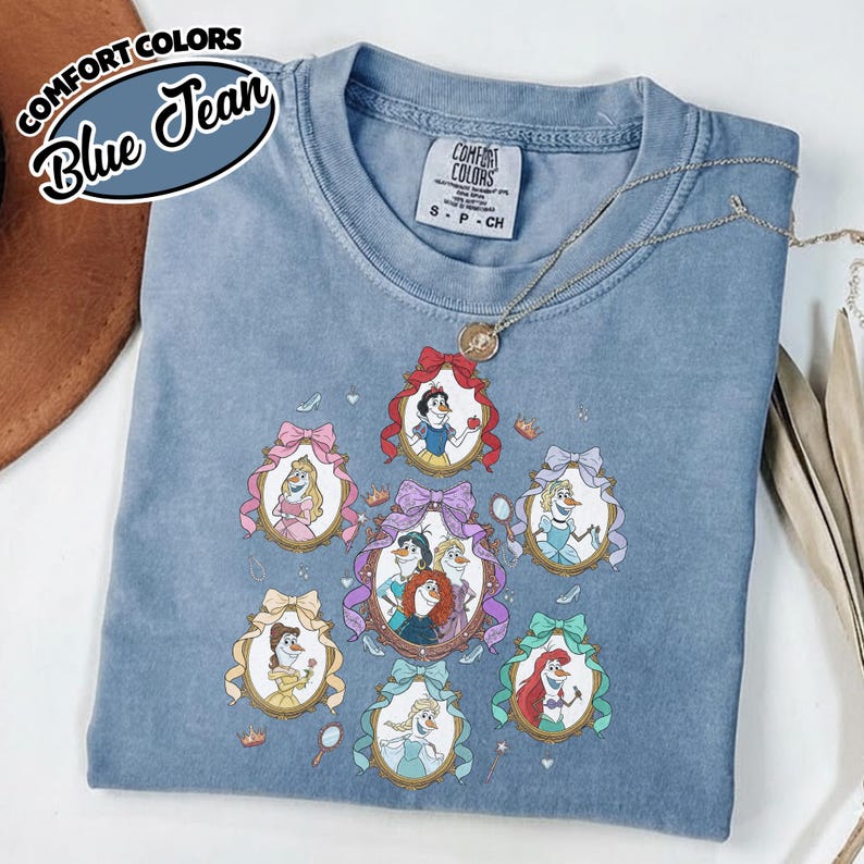 May include: A blue Comfort Colors t-shirt featuring framed illustrations of Disney princesses. The shirt has a crew neck and short sleeves. The text "Comfort Colors Blue Jean" is printed on the upper left corner. A silver necklace is draped across the shirt.