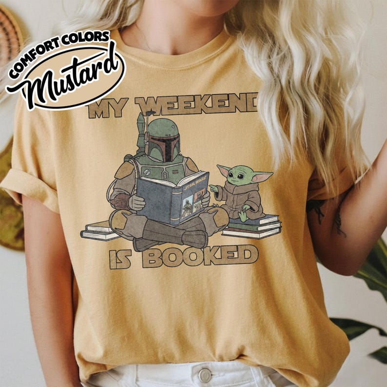Może przedstawiać: Musztardowa koszulka z napisem "MY WEEKEND IS BOOKED". Grafika przedstawia Boba Fetta czytającego książkę "Star Wars" z Grogu. Koszulka ma podwinięte rękawy i etykietę Comfort Colors.