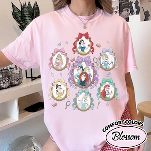 May include: Pink Comfort Colors t-shirt featuring a design of cartoon princesses in ornate frames with bows. The shirt includes the text "Blossom" in a circular design. Accessories like crowns, mirrors, and shoes are also included in the design.