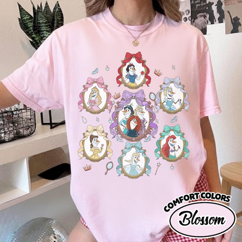 May include: Pink Comfort Colors t-shirt featuring a design of cartoon princesses in ornate frames with bows. The shirt includes the text "Blossom" in a circular design. Accessories like crowns, mirrors, and shoes are also included in the design.