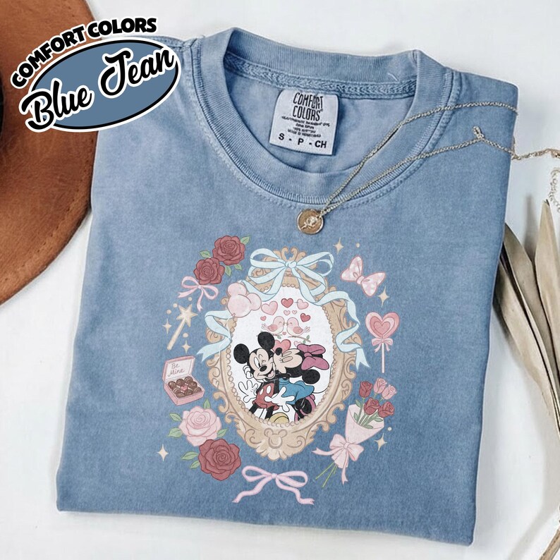 Puede incluir: Una camiseta color azul jean con un gr&aacute;fico de Mickey y Minnie Mouse dentro de un marco ovalado ornamentado. El dise&ntilde;o incluye rosas rojas, una caja de bombones con la etiqueta Be Mine, una piruleta en forma de coraz&oacute;n y un lazo azul.