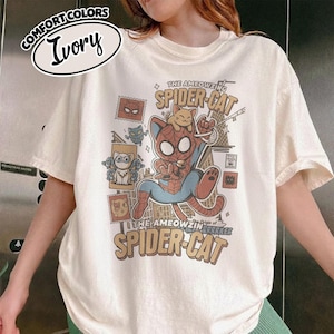 May include: Ivory-colored t-shirt with a graphic of a cat dressed as Spider-Man, with the text "The Ameowzing Spider-Cat" and "The Ameowzing Origin of Spider-Cat". The shirt also features cat-themed postage stamps.