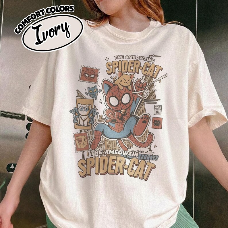 May include: Ivory-colored t-shirt with a graphic of a cat dressed as Spider-Man, with the text "The Ameowzing Spider-Cat" and "The Ameowzing Origin of Spider-Cat". The shirt also features cat-themed postage stamps.