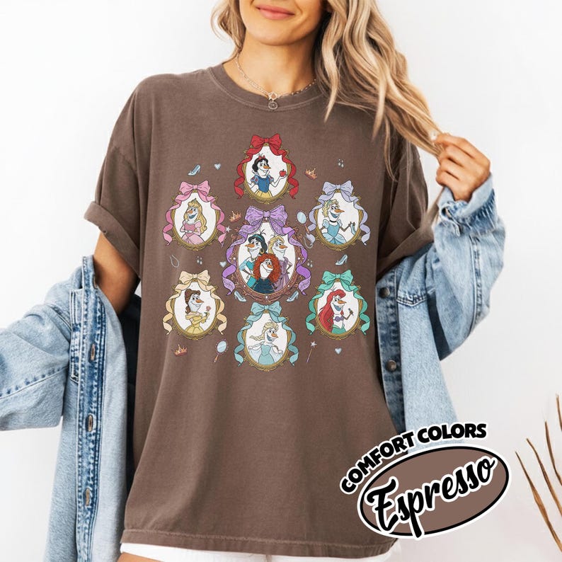 May include: Brown t-shirt featuring framed portraits of animated princesses. Each portrait is surrounded by a decorative bow. The shirt has the words "Comfort Colors Espresso" printed on the bottom right corner.