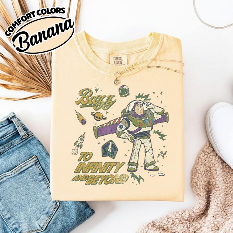 May include: A pale yellow crew neck t-shirt featuring a graphic of the character Buzz Lightyear saluting. The design includes space-themed elements like a rocket, planet, and comet, with the text "Buzz TO INFINITY AND BEYOND" in a retro font.