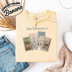 Op de afbeelding: Een lichtgeel T-shirt met de tekst "Walt Disney World" en vier postzegelachtige afbeeldingen van Disney World-attracties. Het shirt heeft de woorden "Comfort Colors" en "Banana" in een cirkelvormig ontwerp. Het shirt wordt gecombineerd met een spijkerbroek en een ketting.
