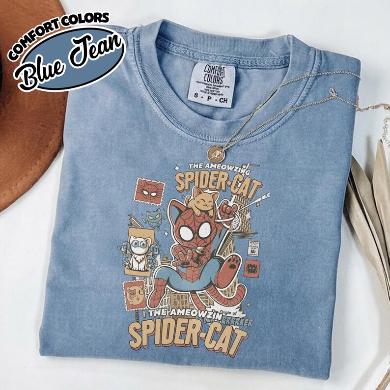 May include: Blue jean Comfort Colors t-shirt featuring a graphic of Spider-Cat. The design includes the text "The Ameowzing Spider-Cat" and various cartoon cat illustrations. A gold necklace is draped across the shirt.