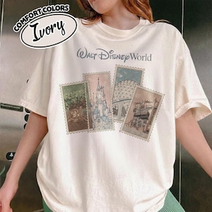 Op de afbeelding: Ivoor kleurig T-shirt met de tekst "Walt Disney World" in een lichtblauwe letter. Het shirt heeft vier postzegel-achtige afbeeldingen van Disney World attracties. De linkerbovenhoek van het shirt heeft een Comfort Colors logo met het woord "Ivory".