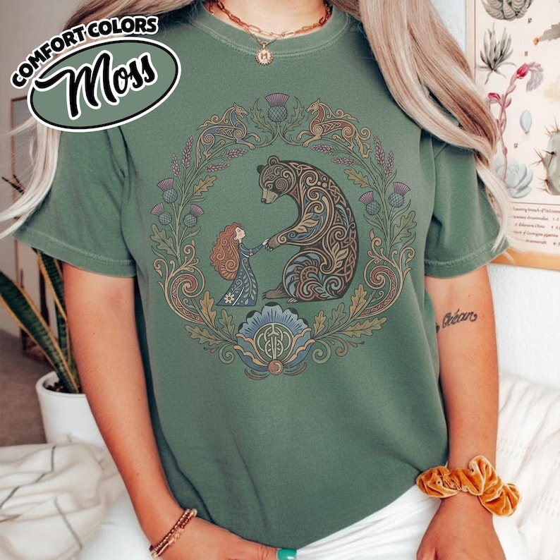 May include: Moss green t-shirt featuring a circular design with a bear and a girl holding hands, surrounded by floral and Celtic knot elements. The shirt has the words "COMFORT COLORS" and "Moss" in a green oval on the upper left.