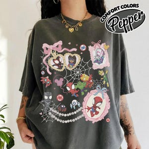 K&ouml;nnte beinhalten: Dunkelgraues T-Shirt mit einer farbenfrohen Grafik verschiedener Spider-Man-Charaktere in rosa Rahmen und einem Spinnennetz-Design. Das Shirt hat den Text "COMFORT COLORS Pepper" in einem kreisf&ouml;rmigen Design.