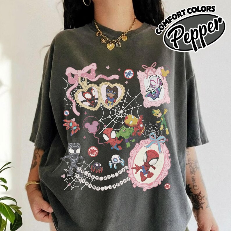 K&ouml;nnte beinhalten: Dunkelgraues T-Shirt mit einer farbenfrohen Grafik verschiedener Spider-Man-Charaktere in rosa Rahmen und einem Spinnennetz-Design. Das Shirt hat den Text "COMFORT COLORS Pepper" in einem kreisf&ouml;rmigen Design.