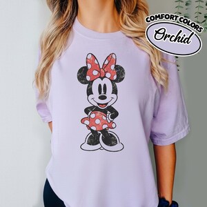 May include: Lilac t-shirt with a distressed Minnie Mouse graphic, sporting a red polka-dot bow and dress. The shirt has a vintage aesthetic, with the words "COMFORT COLORS" and "Orchid" printed on the upper right.