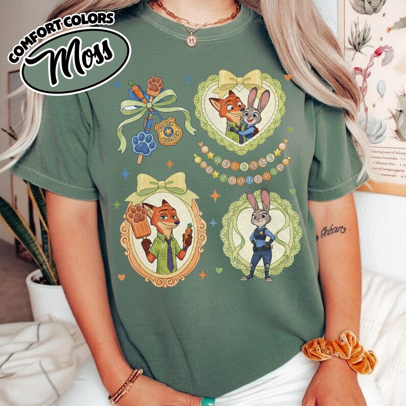 May include: A moss green crewneck t-shirt featuring Zootopia character graphics. The design includes Nick Wilde and Judy Hopps in ornate frames, a police badge, a paw print, and the text Welcome to Zootopia. The shirt has a Comfort Colors label.