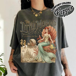 以下が含まれることがあります： ヴィンテージイラストが特徴のダークグレーのTシャツ。人魚姫のアリエルが貝殻の玉座に座り、城、タツノオトシゴ、テキスト「A Vintage Sea Tale」が描かれています。半袖でゆったりとしたフィット感です。