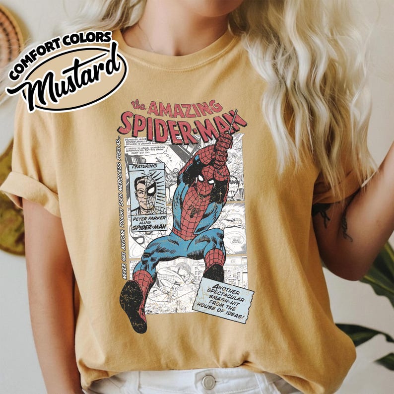 K&ouml;nnte beinhalten: Senffarbenes T-Shirt mit einer Comic-Grafik von Spider-Man. Das Design enth&auml;lt den Text "the AMAZING SPIDER-MAN" und andere Comic-Elemente. Das T-Shirt ist mit "COMFORT COLORS" gekennzeichnet.