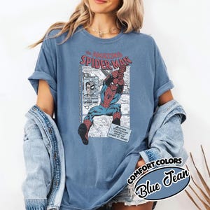 K&ouml;nnte beinhalten: Blaues Jeans Comfort Colors T-Shirt mit einem Vintage-Comic-Grafik von Spider-Man. Die Grafik enth&auml;lt den Text "The Amazing Spider-Man" und Comic-Panels. Das Shirt hat hochgekrempelte &Auml;rmel.