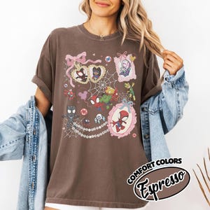 K&ouml;nnte beinhalten: Espressofarbenes T-Shirt mit einer farbenfrohen Grafik von Spider-Man-Charakteren in verzierten Rahmen, die durch ein Spinnennetzdesign verbunden sind. Das Shirt hat hochgekrempelte &Auml;rmel und den Text "COMFORT COLORS Espresso" in einem kreisf&ouml;rmigen Design.