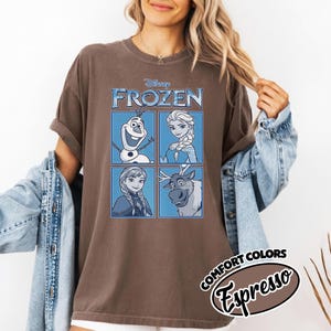 May include: Brown t-shirt featuring the Disney movie "Frozen." The shirt displays four character portraits in blue squares: Olaf, Elsa, Anna, and Sven. The text "FROZEN" is above the portraits. The shirt also has the text "COMFORT COLORS Espresso."