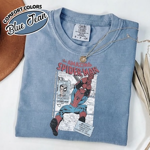 K&ouml;nnte beinhalten: Blaues Comfort Colors T-Shirt mit einem Spider-Man-Motiv aus der Comic-Serie. Das Shirt zeigt den Text "the AMAZING SPIDER-MAN" und Comic-Panels. Eine silberne Halskette mit Anh&auml;nger liegt auf dem Shirt.