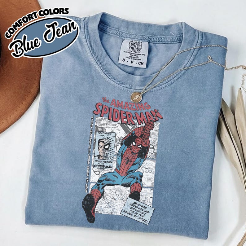K&ouml;nnte beinhalten: Blaues Comfort Colors T-Shirt mit einem Spider-Man-Motiv aus der Comic-Serie. Das Shirt zeigt den Text "the AMAZING SPIDER-MAN" und Comic-Panels. Eine silberne Halskette mit Anh&auml;nger liegt auf dem Shirt.