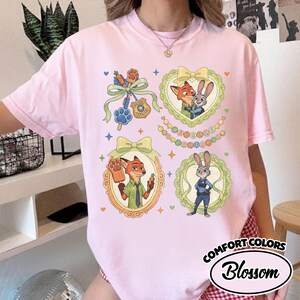 May include: A light pink t-shirt featuring a graphic print of Zootopia characters Nick Wilde and Judy Hopps. The design includes ornate frames, bows, a police badge, a paw-shaped popsicle, and the text Welcome to Zootopia in colorful circles.