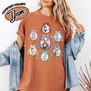 May include: Rust-colored t-shirt featuring a graphic of several princesses in ornate frames with bows. The shirt has the words "Comfort Colors" and "Yam" in a brown oval on the upper left.